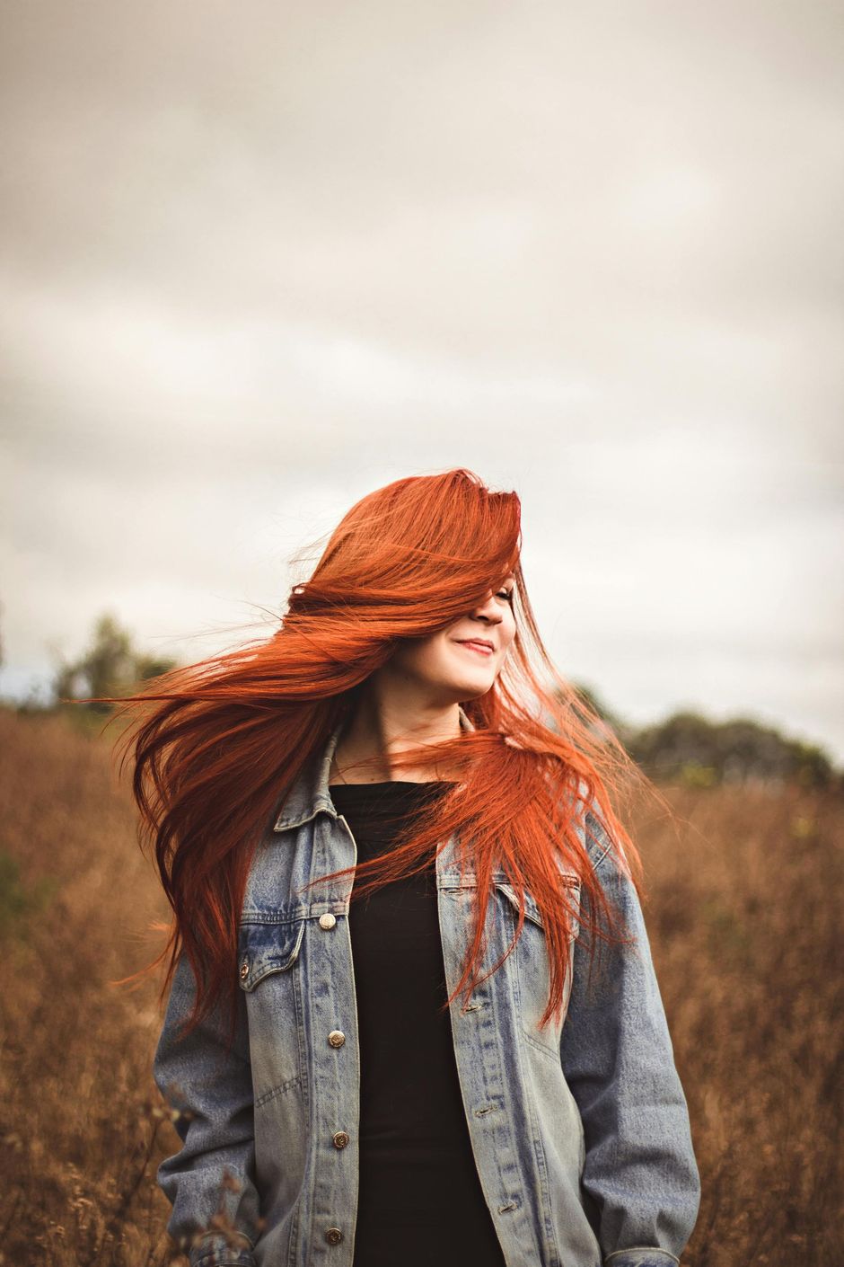 Person with long red hair in a denim jacket standing in a field, looking to the side