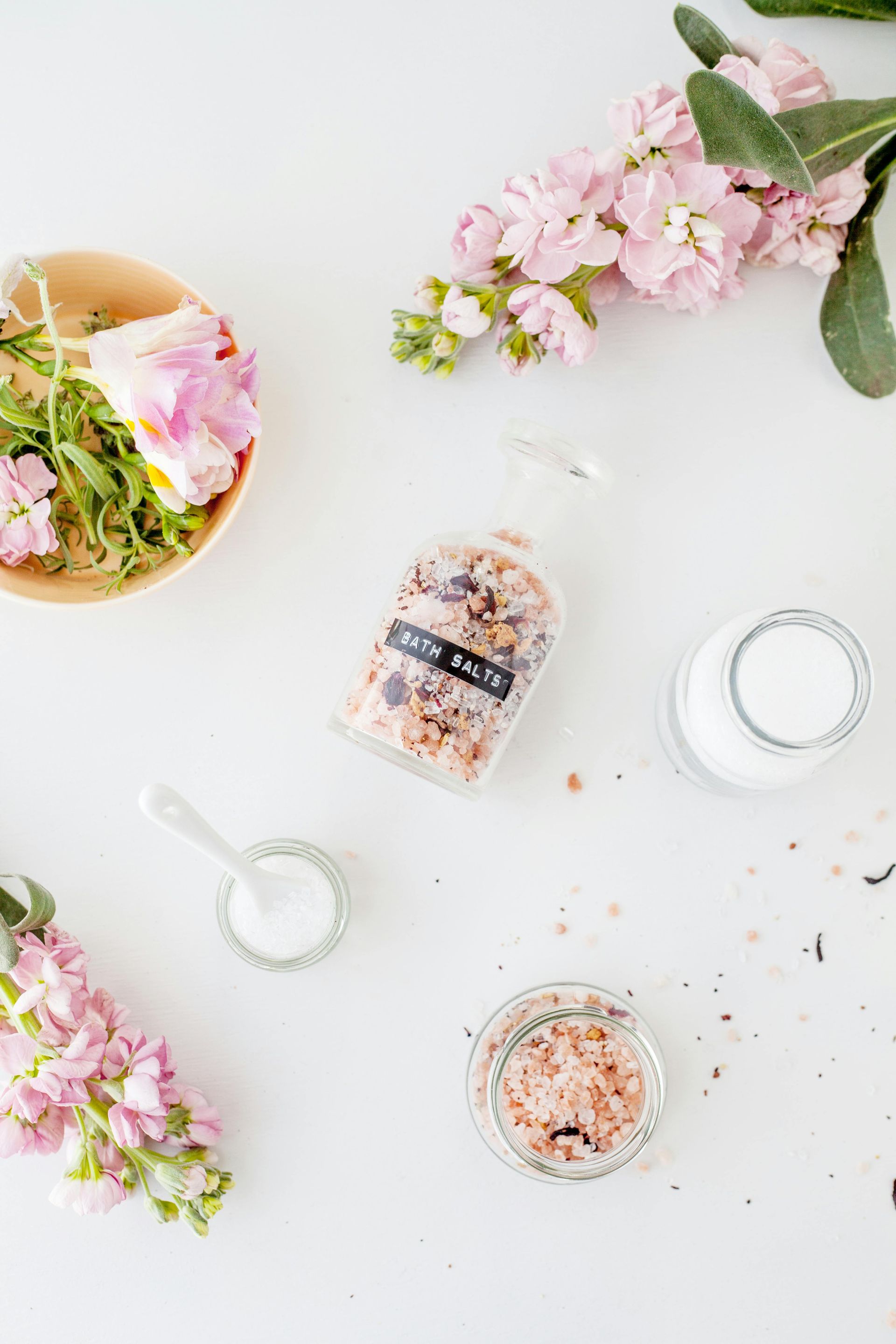 Flat lay of pastel flowers, bath salts, jars, and white candles on a white background