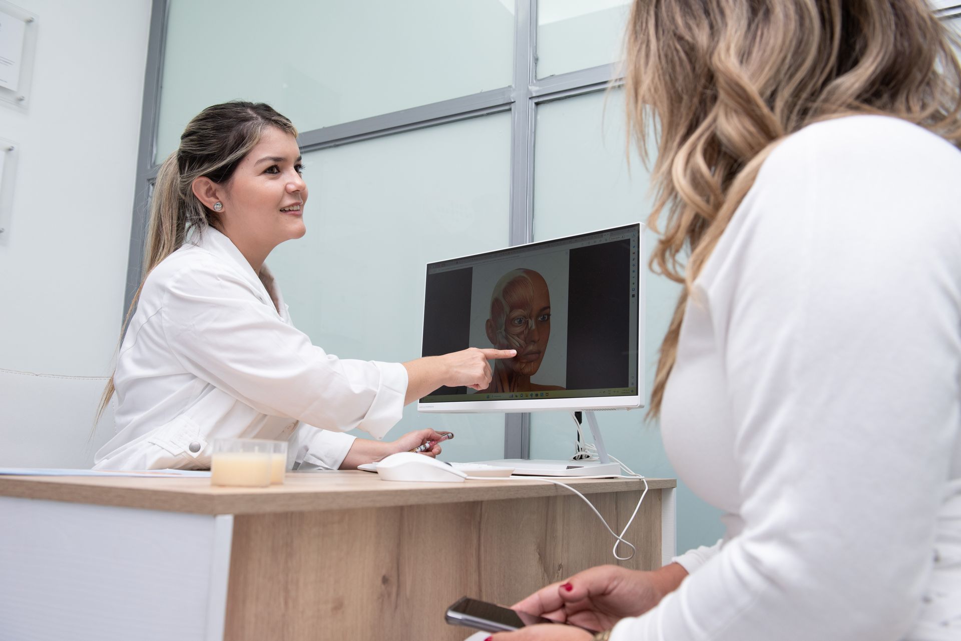 Una mujer está sentada en un escritorio hablando con un médico que señala la pantalla de una computadora.