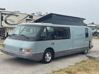 Light blue and gray RV with a pop-up roof parked outside, RV park in the background.