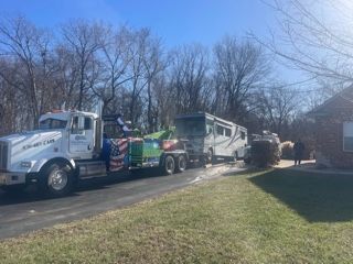 A tow truck pulling a damaged RV from a driveway on a sunny day.