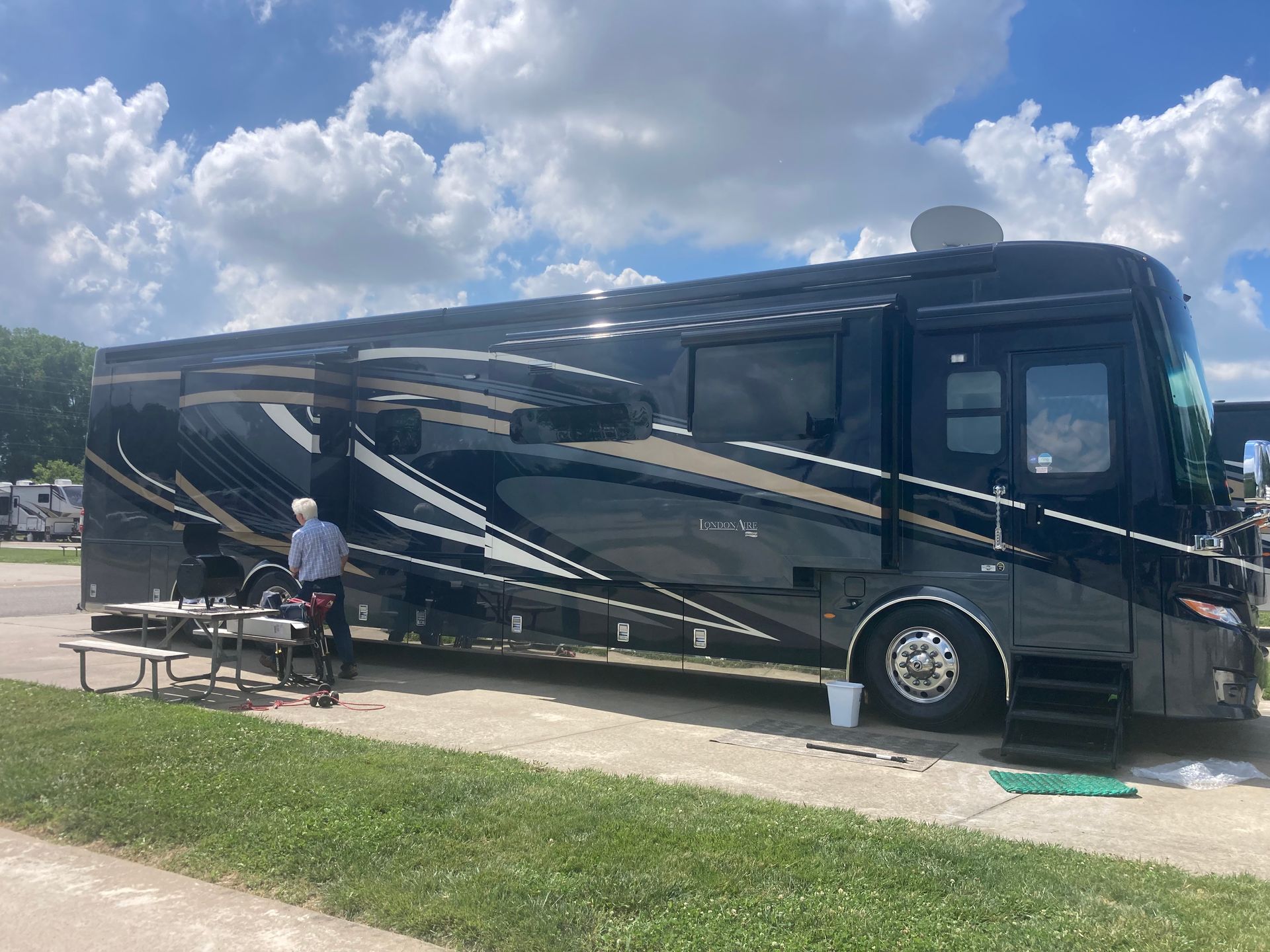 Black RV parked outdoors, person standing near a picnic table on a sunny day.