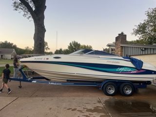White and teal boat on a blue trailer parked on a driveway, two people nearby.