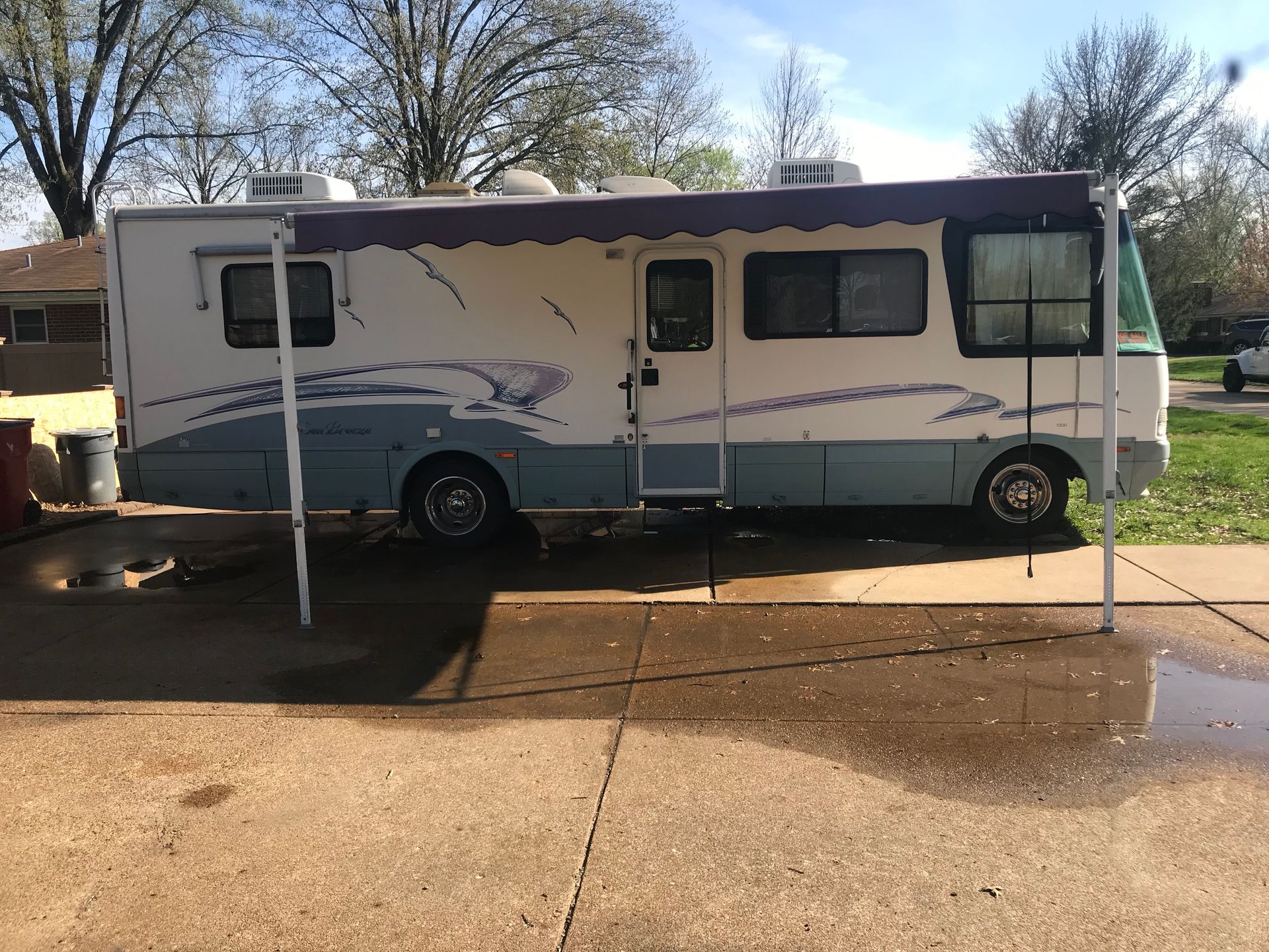 RV parked on a concrete driveway, two-tone paint: light blue and white with purple awning, door, windows, and awning support.