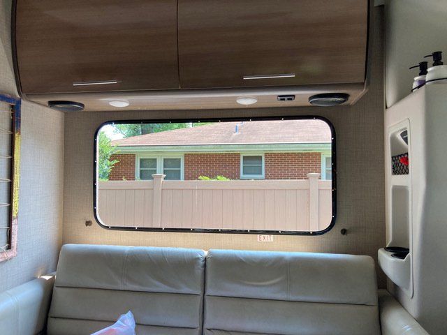 Interior view of a campervan with a window overlooking a fenced yard and brick house. Beige upholstery and cabinets.