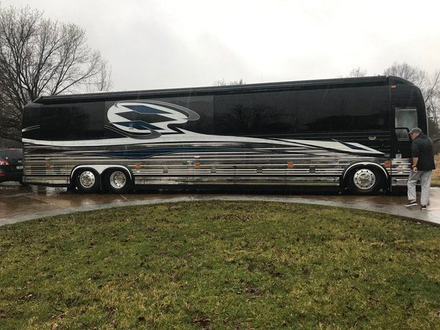 Black and chrome luxury bus parked on a wet, circular driveway. A person stands nearby.