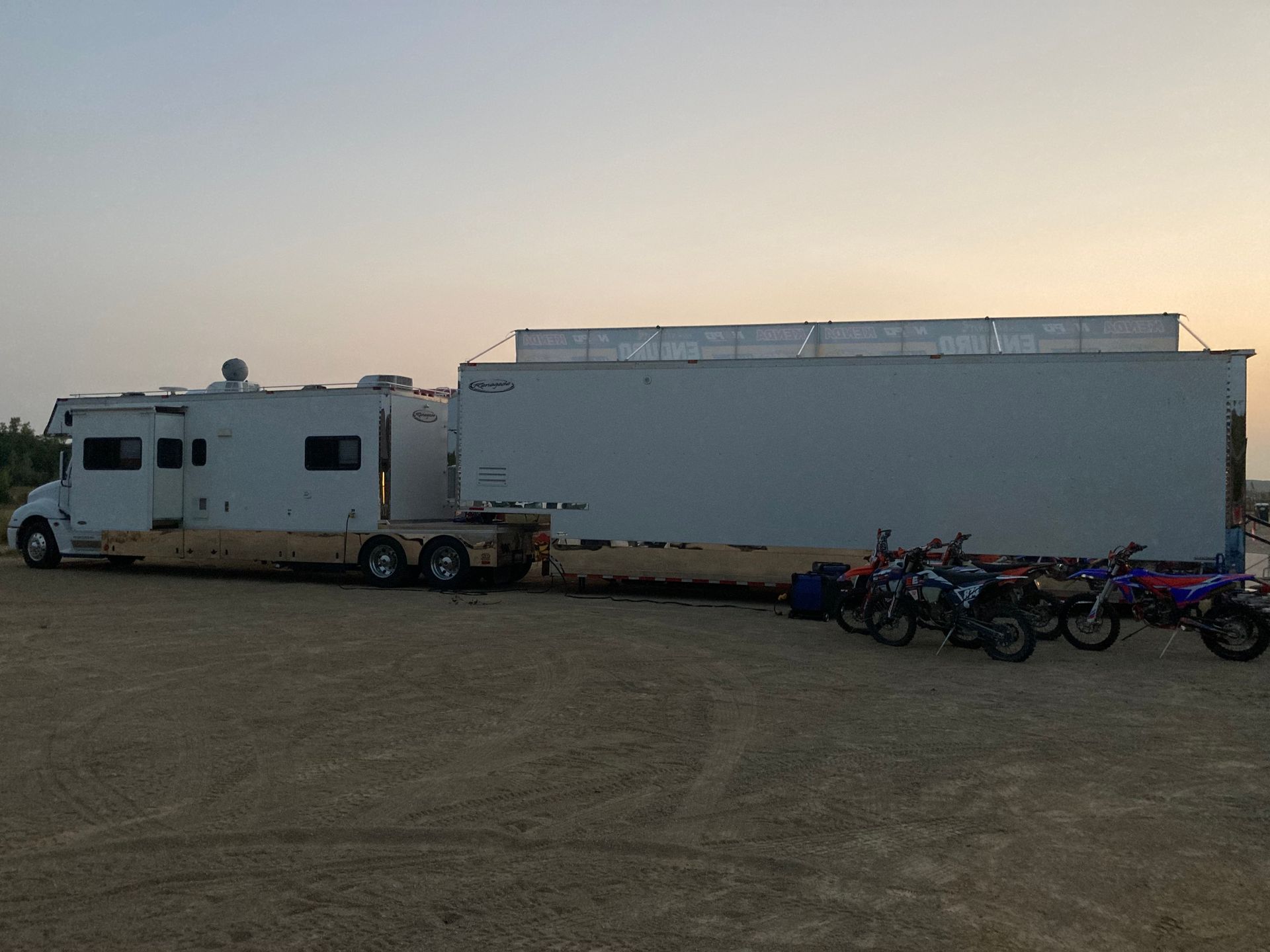 White semi-truck with trailer, dirt bikes parked nearby, set against a sunset sky.
