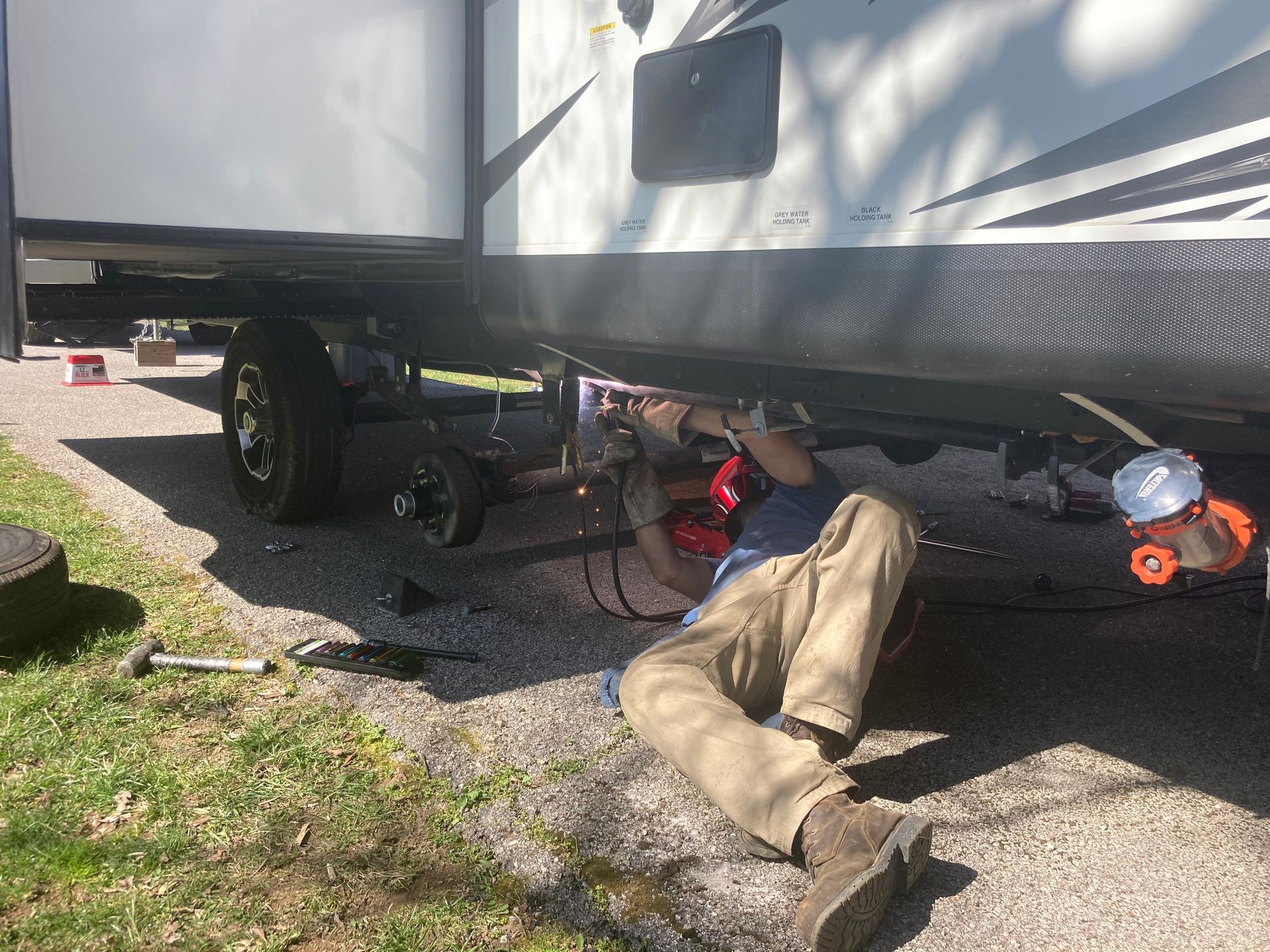 A person working under an RV, holding a tool; outdoors, sunny.