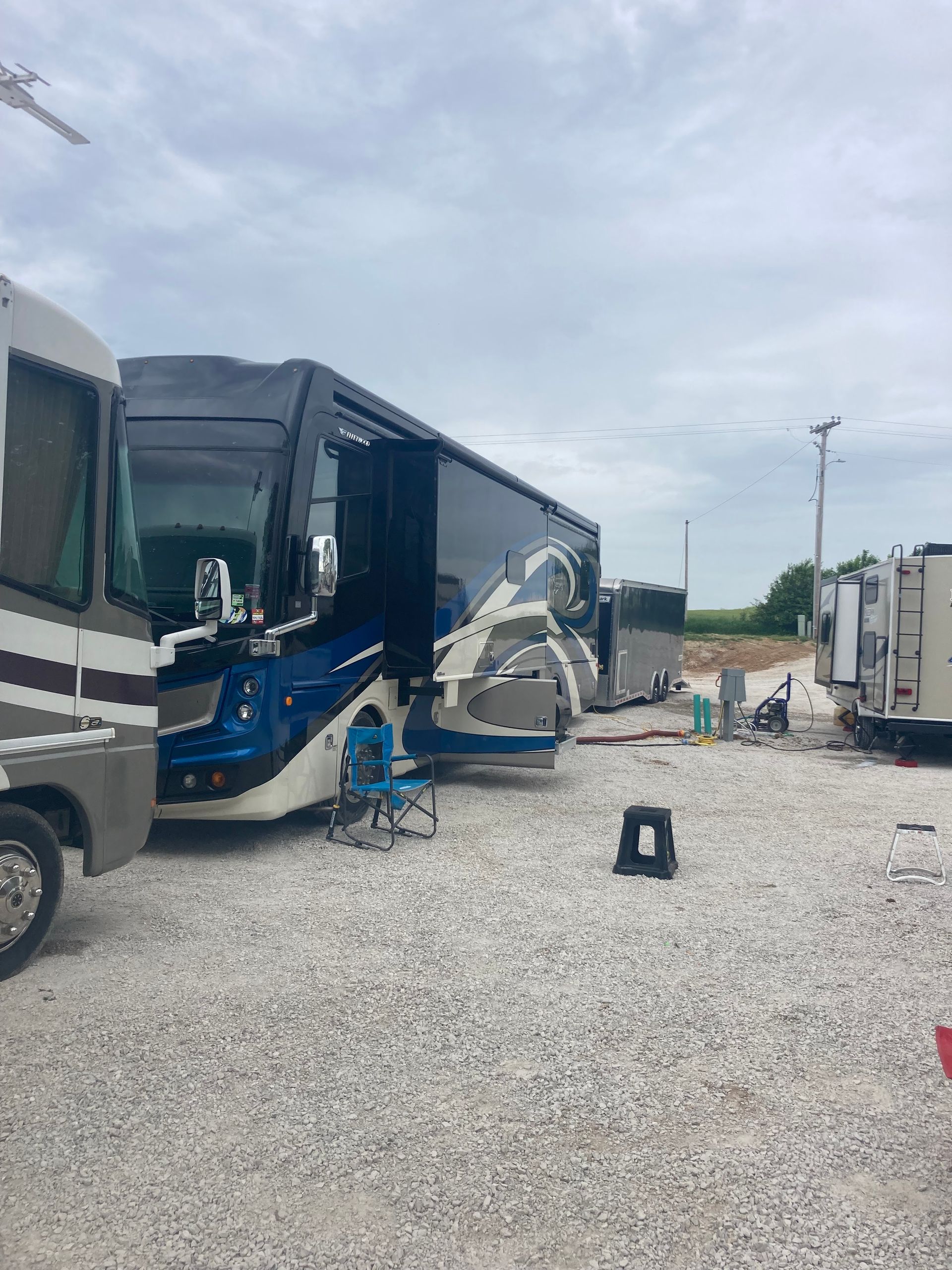 Large RV parked on gravel, awning extended. Blue and white paint. Camp chair and step stool are visible.