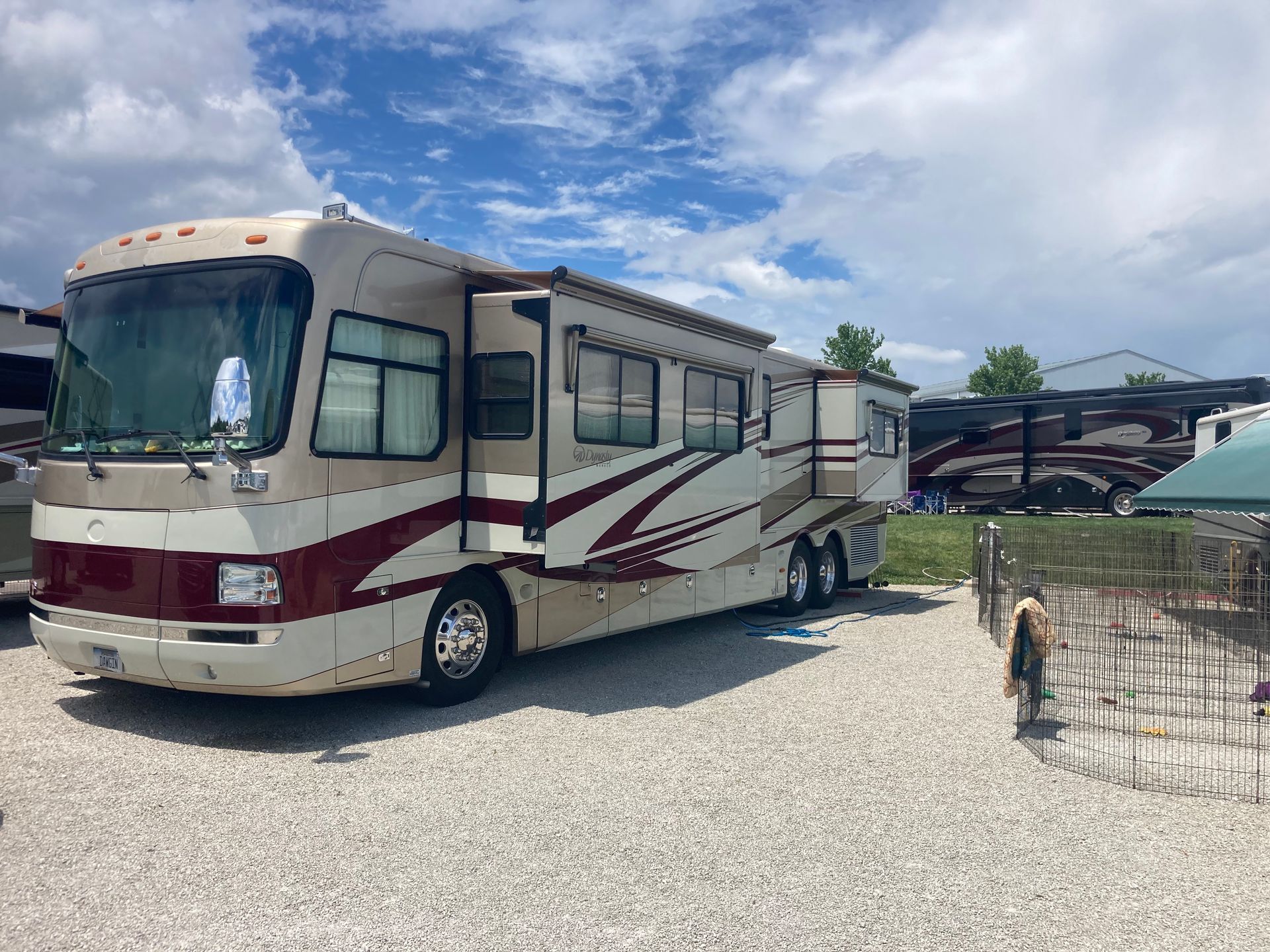 Beige and maroon RV parked at a campsite with its awning extended under a cloudy sky.