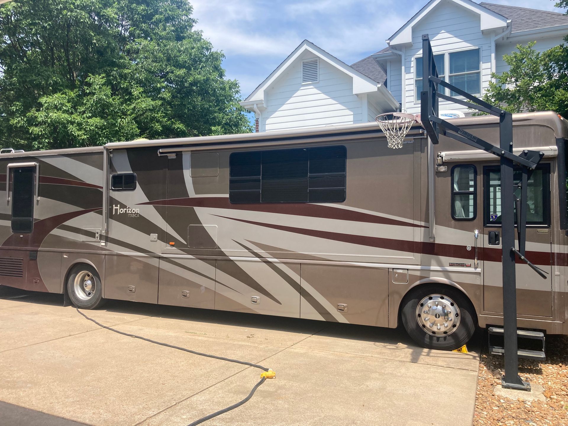 A tan RV parked in a driveway next to a basketball hoop and a two-story house.