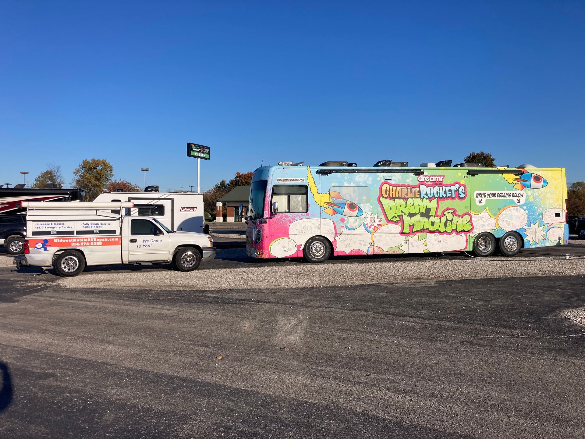 A colorful, cartoon-decorated tour bus parked next to a white pickup truck in a gravel lot under a blue sky.