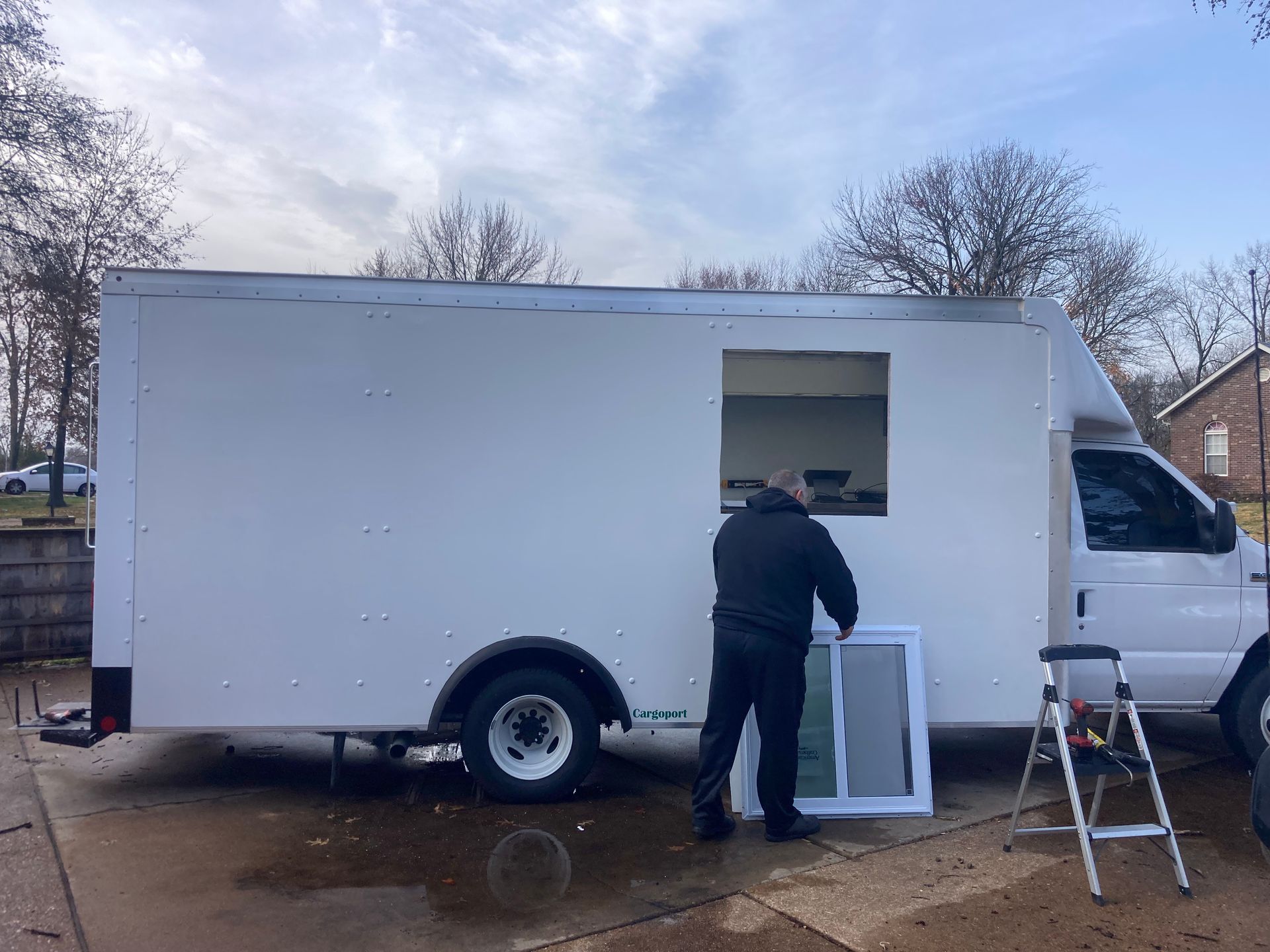 Person installing a window on the side of a white box truck. The truck is parked outdoors on a cloudy day.