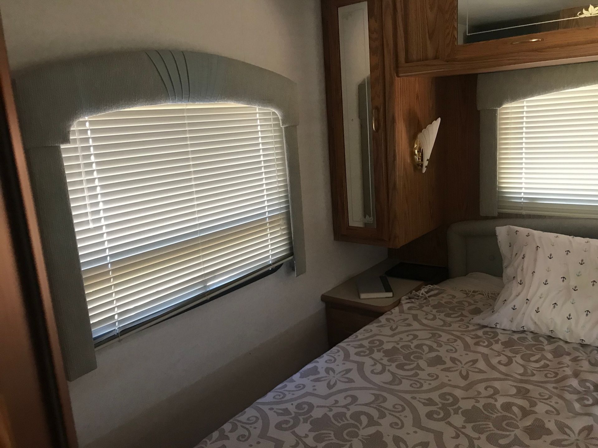 Bedroom interior of a recreational vehicle; window with blinds, bed with patterned comforter.