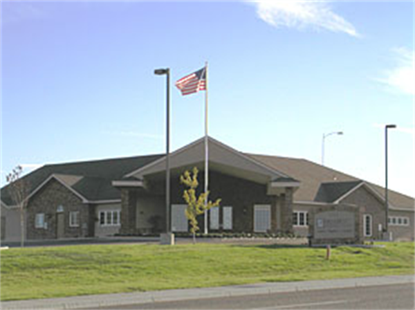 Single-story brick civic building with an American flag, lampposts, and a grassy lawn under a blue sky