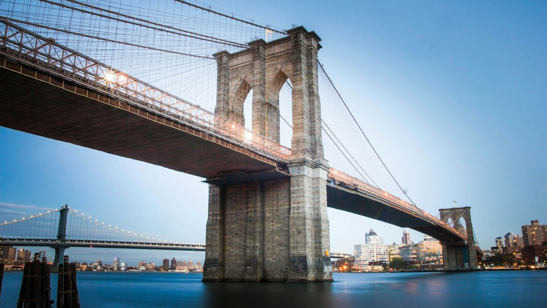 The Brooklyn Bridge towers over the East River with city buildings visible in the background under a blue sky.