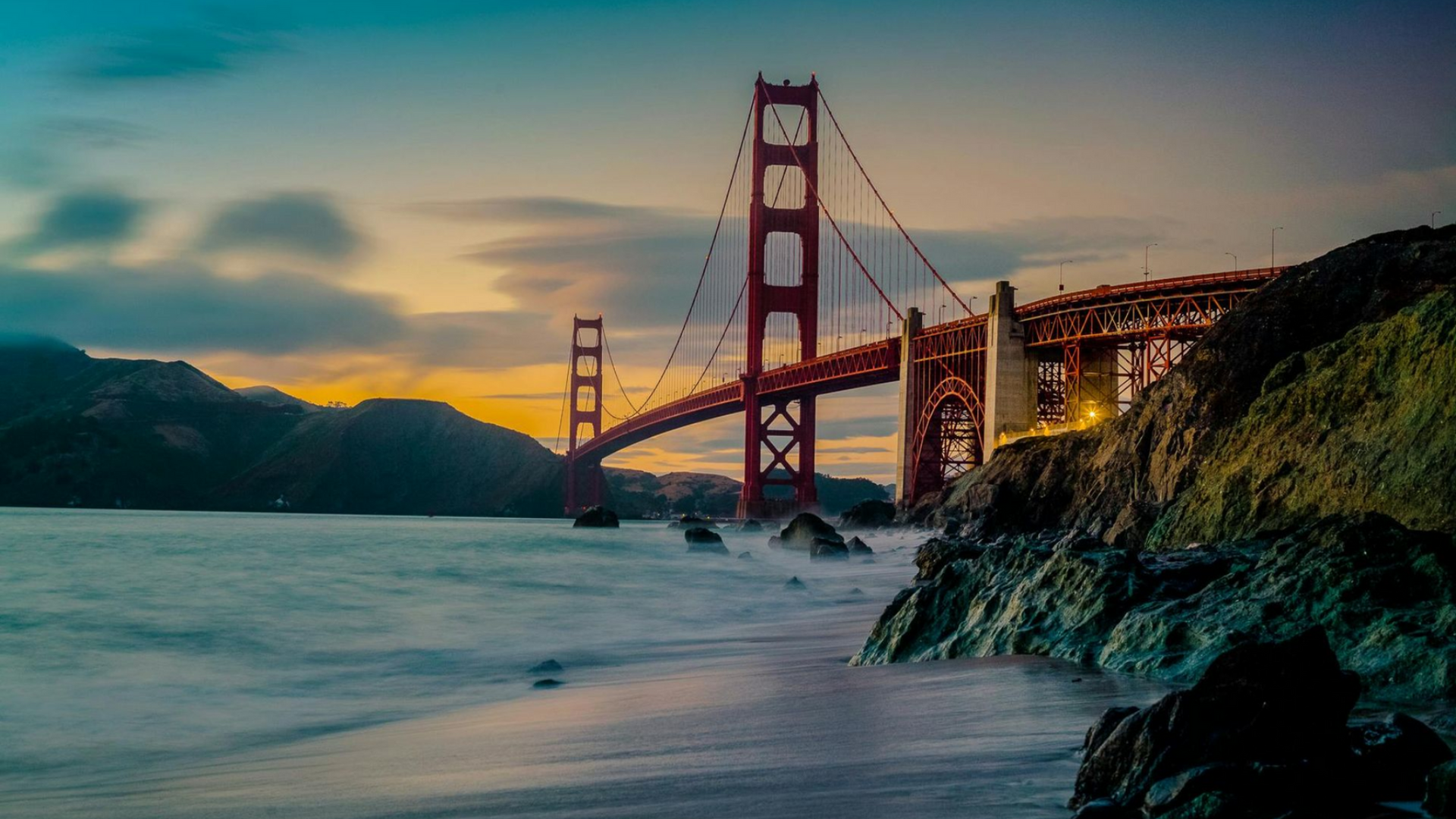 Golden Gate Bridge at sunset with rocky shore and waves in the foreground under a dramatic, colorful sky.