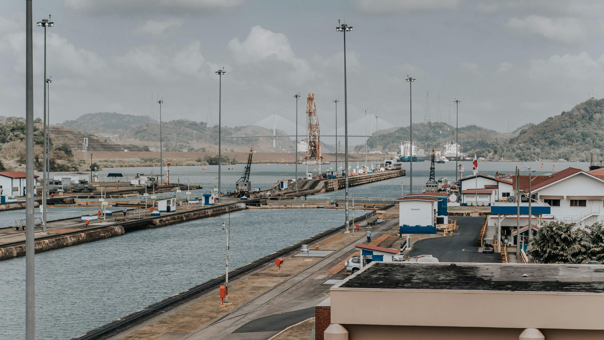 A daytime view of the Panama Canal, showing a narrow waterway, lock gates, industrial infrastructure, and distant hills.