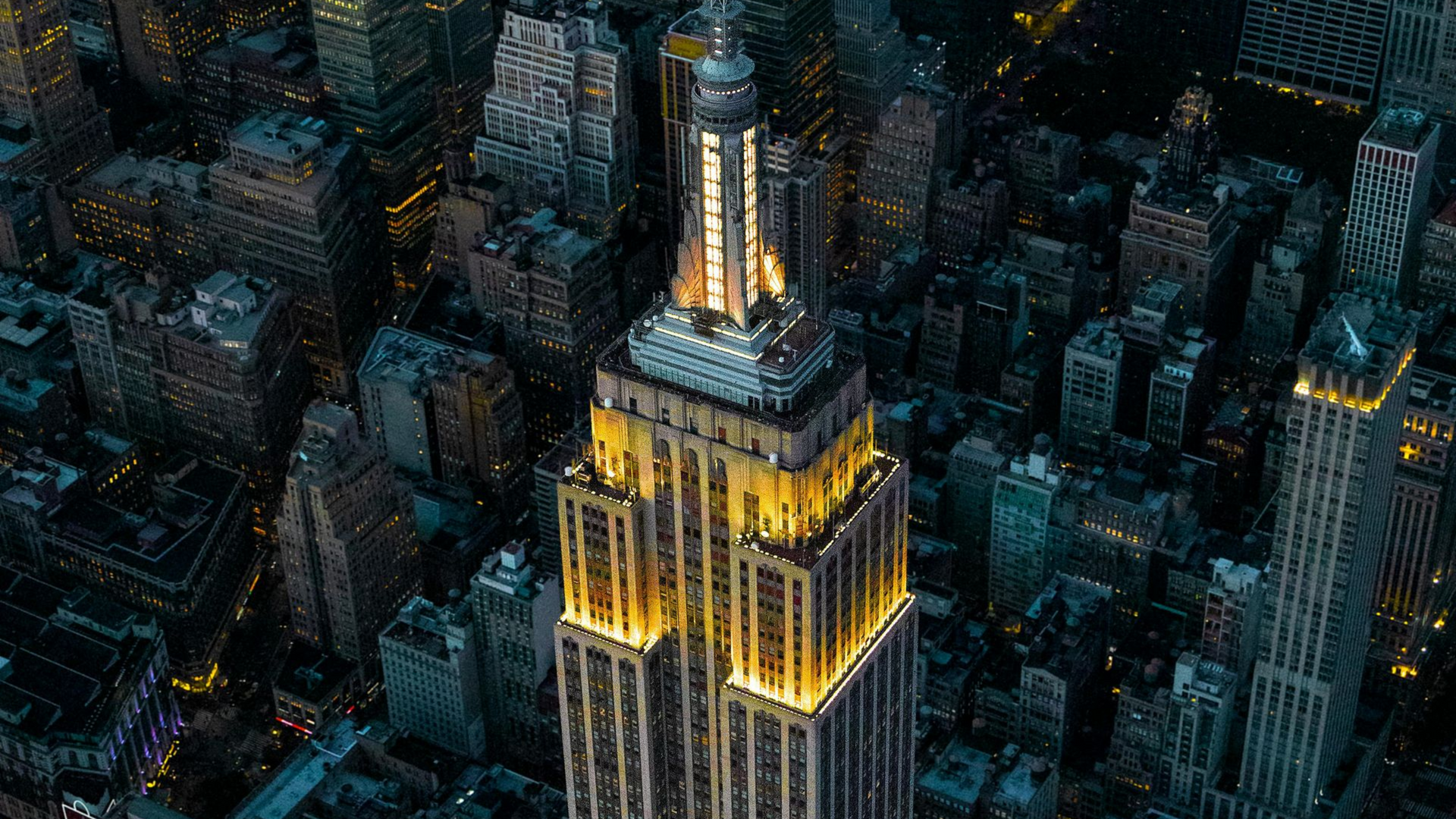 Aerial view of the Empire State Building illuminated with gold lights at night amidst a dense New York City skyline.