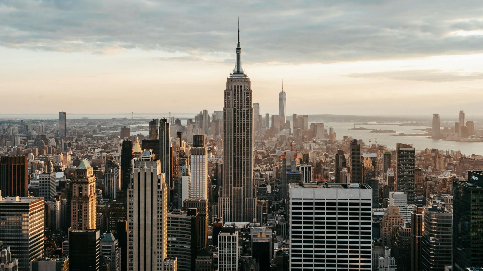 A high-angle view of the Empire State Building towering over the dense, beige-toned skyline of New York City at sunset.