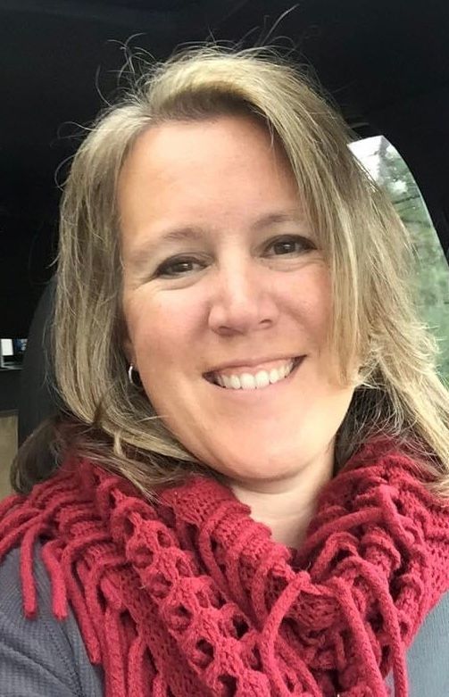 A person with shoulder-length blonde hair smiles while wearing a red knit infinity scarf in a car.