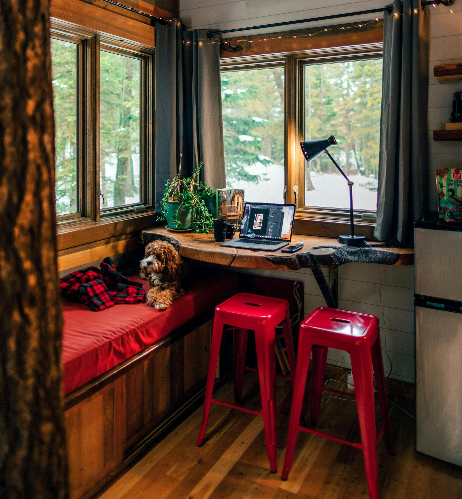A cozy corner office with a window view of snow, featuring a laptop on a wooden desk, two red stools, and a small dog.