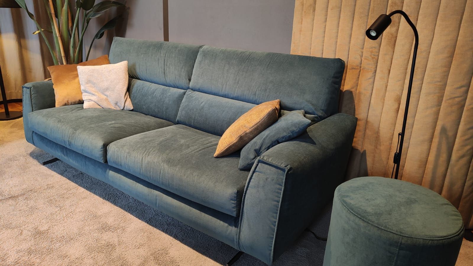 Divano color verde acqua con cuscini, pouf coordinato, lampada e tappeto neutro in una stanza.