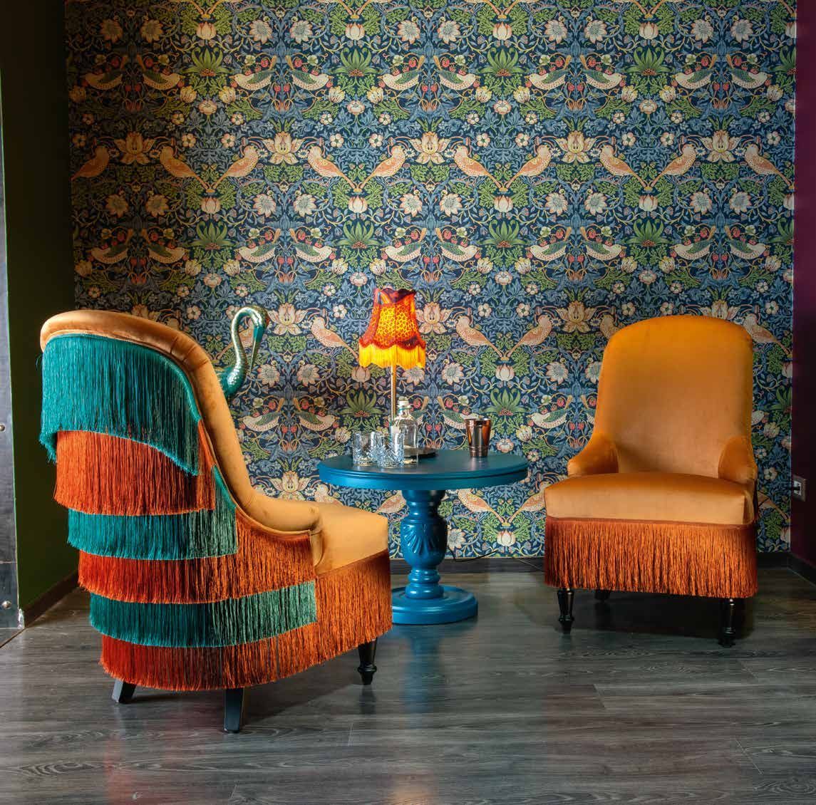 Due sedie arancioni con frange, tavolo blu con lampada, su sfondo di carta da parati floreale.