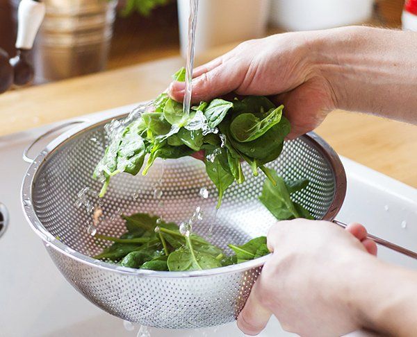 Person washing spinach leaves in a colander under running water in a kitchen sink.