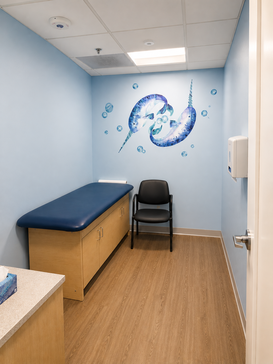 Small exam room with blue walls, exam table, chair, and whale mural.