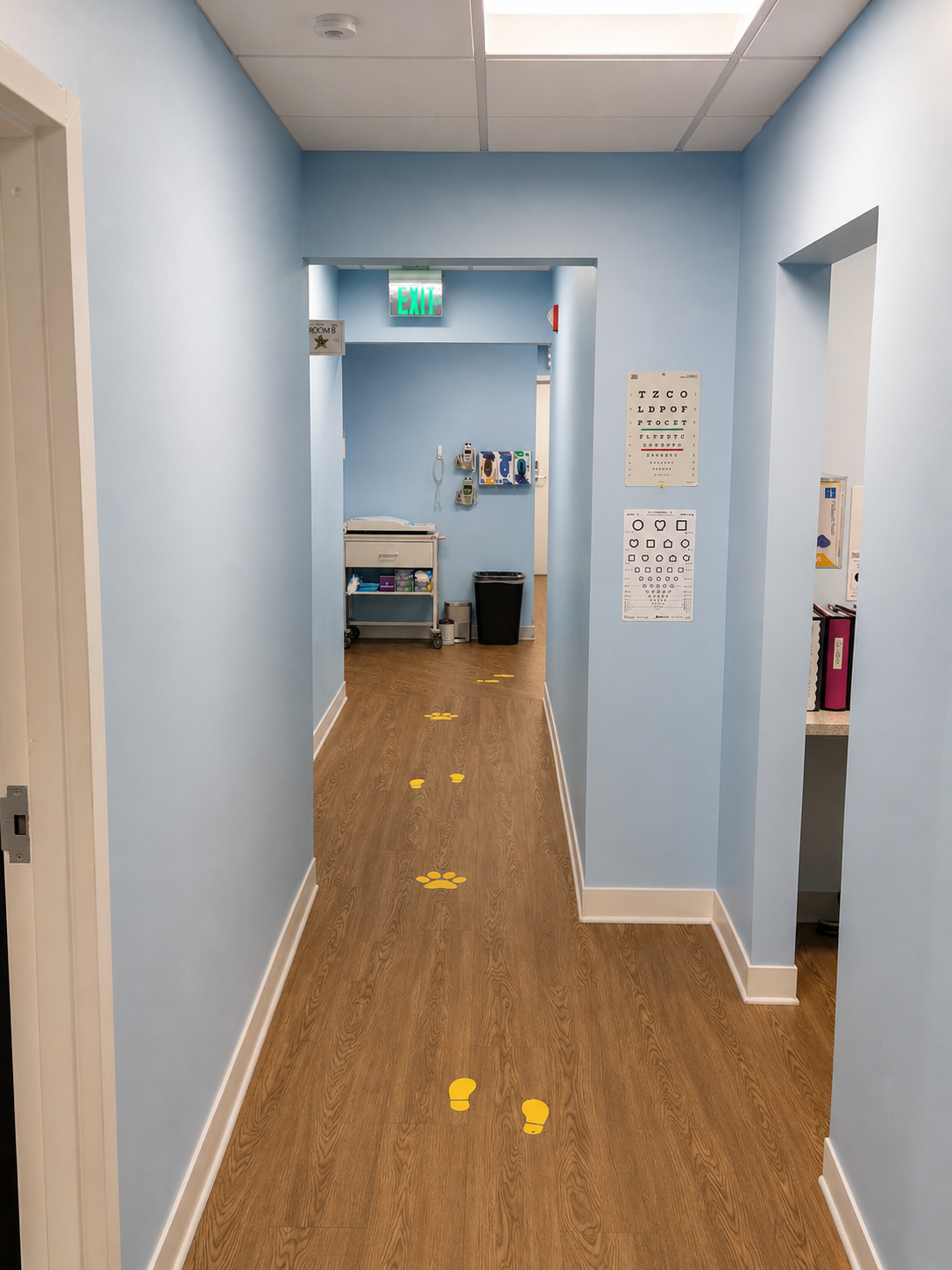 Narrow light-blue hallway with wood floor, yellow floor markers, and an open doorway at the end.