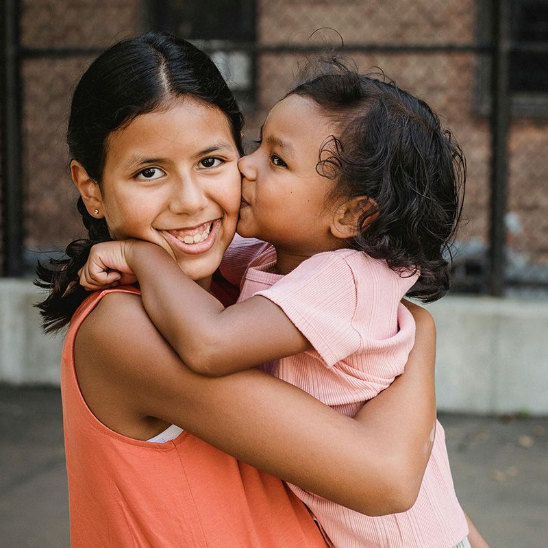 Two children hugging outdoors, one kissing the other’s cheek.