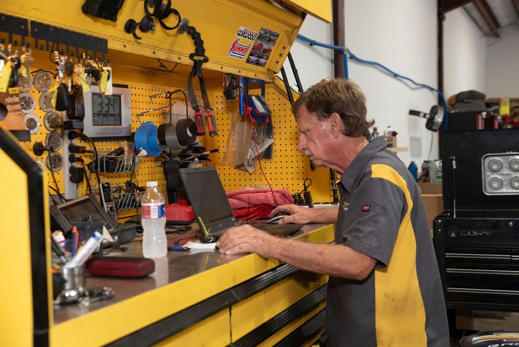Mechanic working at a laptop on a workbench in a yellow tool-filled workshop.