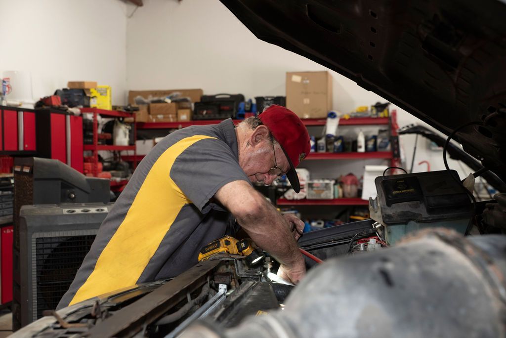 Mechanic in red cap working under a car hood in a garage with shelves of tools behind him