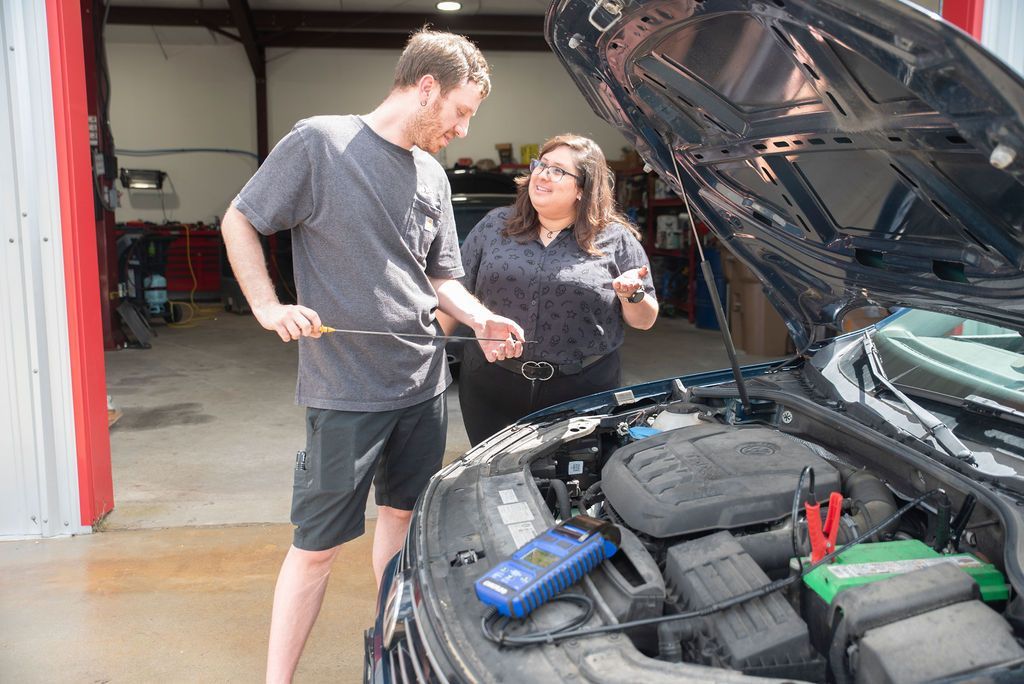 Two people examine a car engine in a garage, with the hood open.
