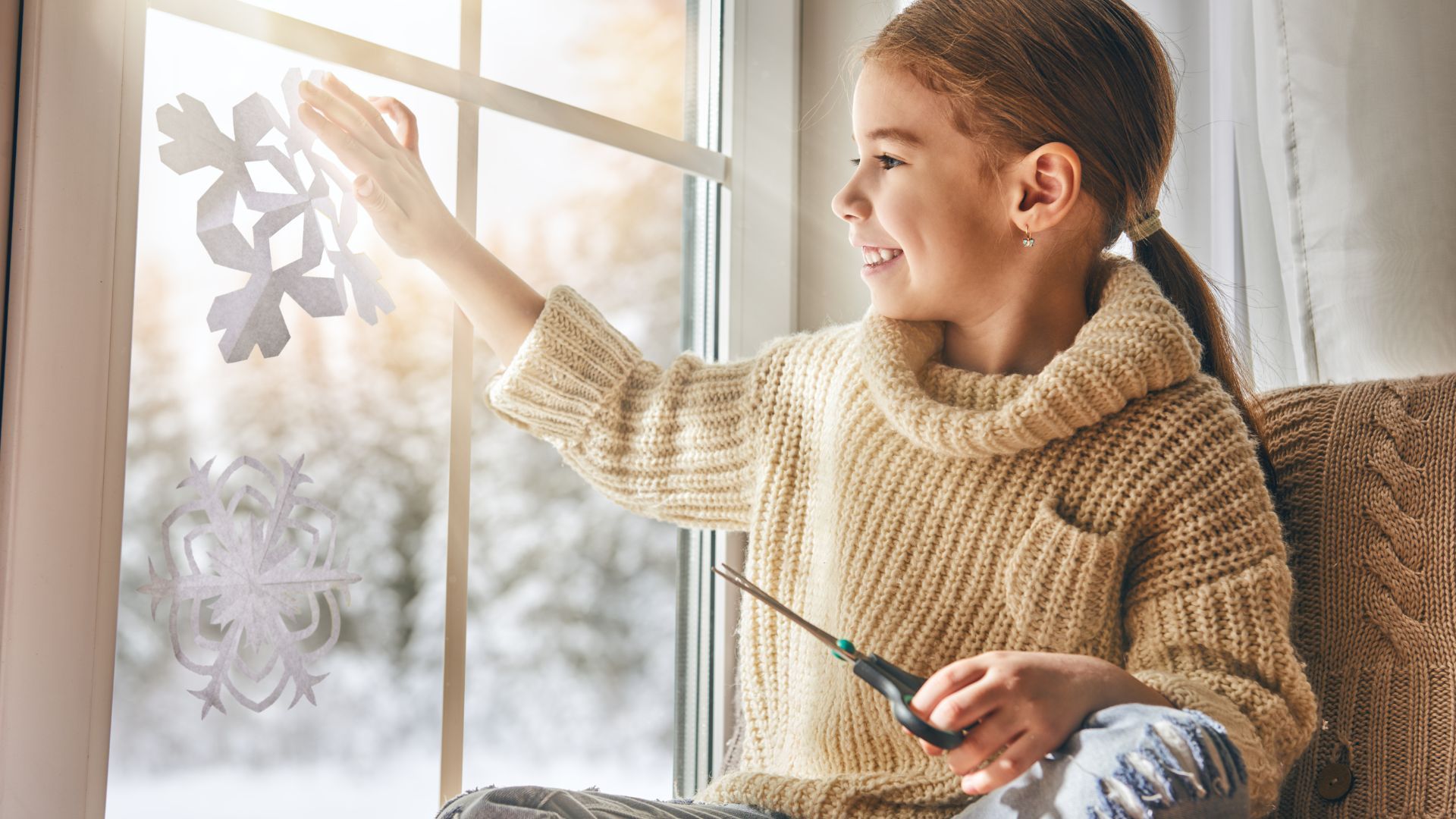 A little girl is sitting on a couch cutting paper snowflakes on a window.