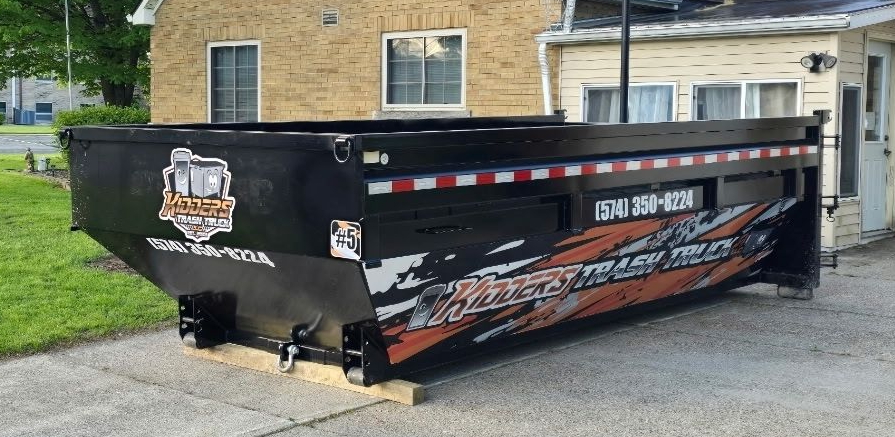 15 Yard Dumpster Black with orange-and-white business signage sitting beside a house and basketball hoop, with a red fire hydrant in front.
