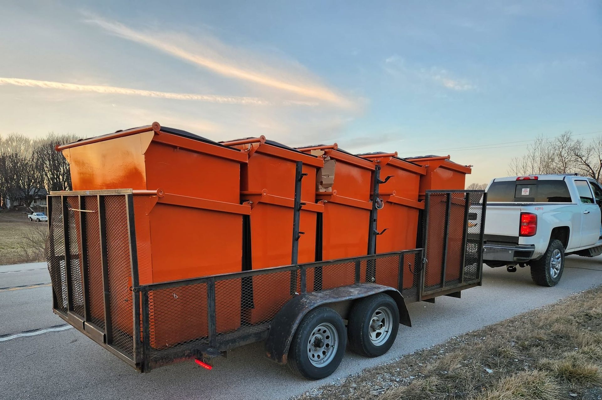 Orange metal cargo trailer loaded with equipment, hitched to a white pickup truck on a roadside at sunset
