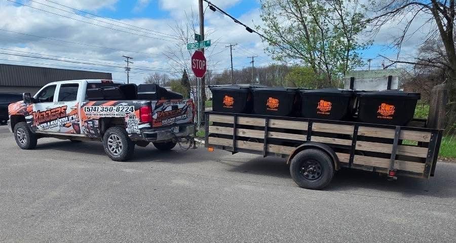 Truck towing a black trailer with orange logos on a street, outdoors near trees and traffic lights