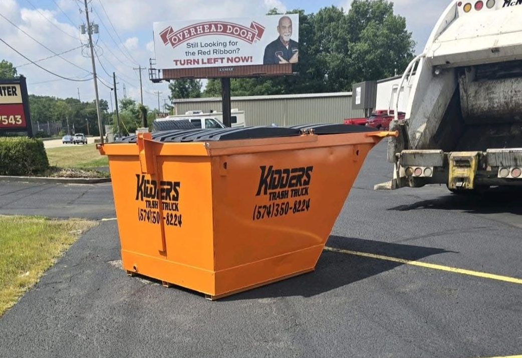Orange construction dumpster in a parking lot beside a garbage truck, with yellow painted lines and a roadside billboard.