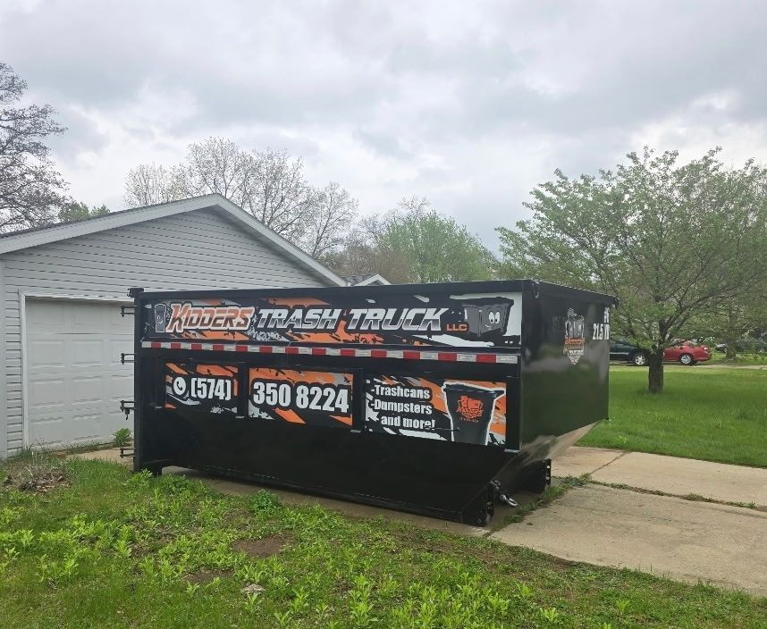 Black dumpster trailer with business ads parked in a driveway beside a gray garage on a cloudy day