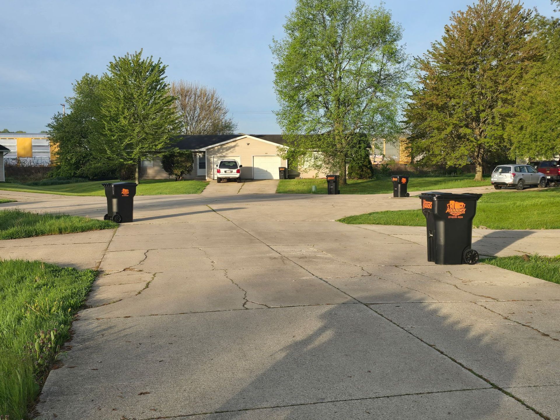 Cracked suburban driveway lined with black trash bins, leading to a house and trees on a sunny day