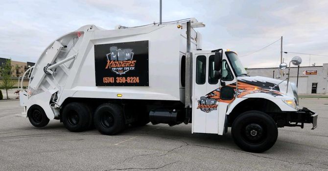 White garbage truck with orange and black logo, parked outdoors on a street under cloudy skies. The Sign on the side of the trash truck reads: Kidder's Trash Truck