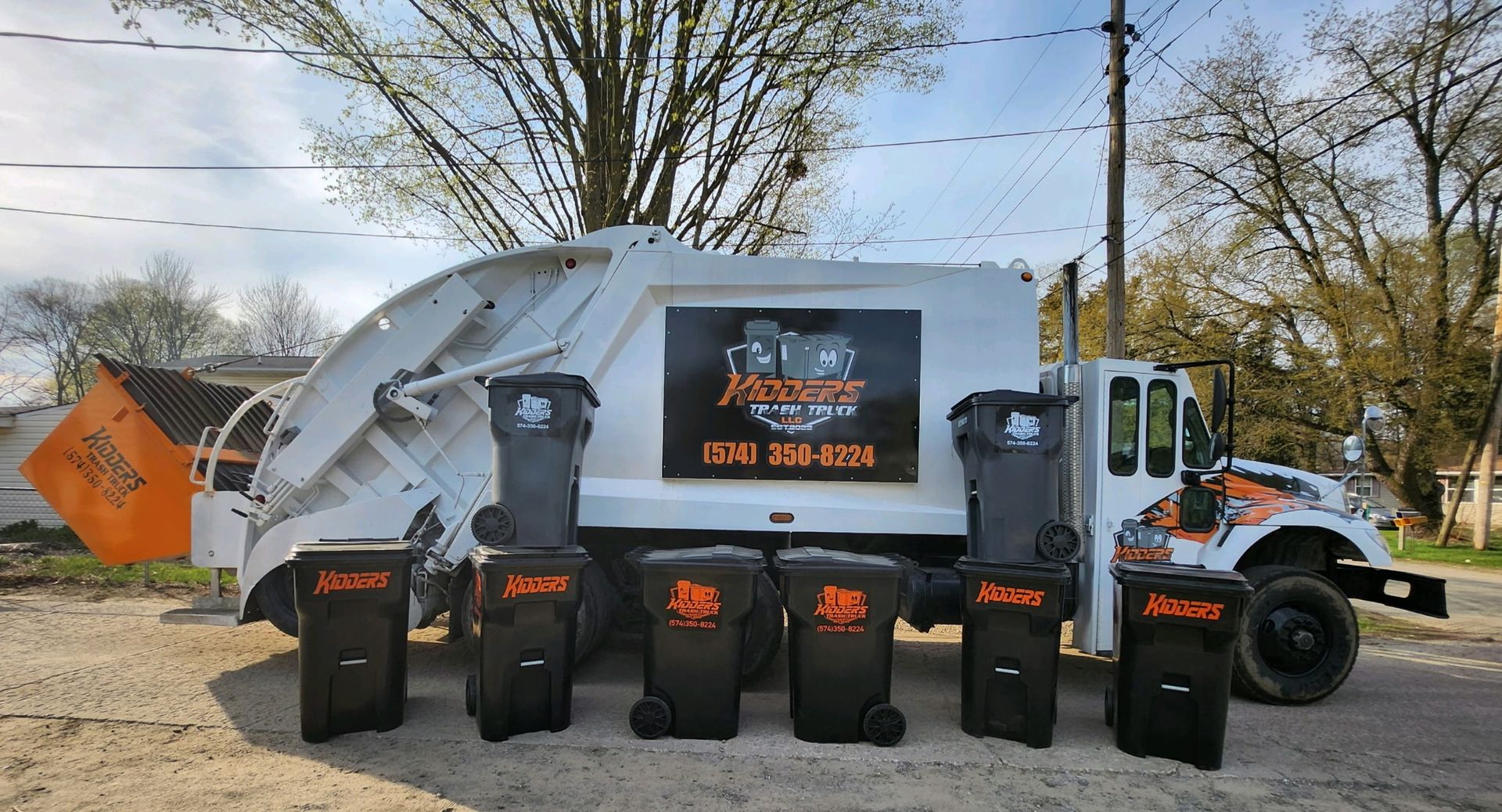 White garbage truck with orange and black logo, parked outdoors on a street under cloudy skies. The Sign on the side of the trash truck reads: Kidder's Trash Truck