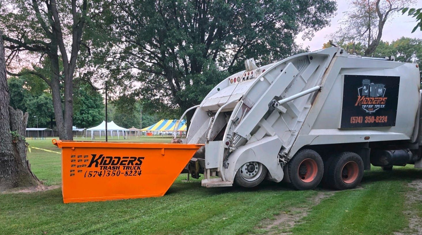 Garbage truck emptying a bright orange Elkhart County dumpster in a grassy park area for commercial use