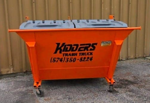 Orange Kidders trash truck dumpster with gray lid and black wheels on pavement outdoors