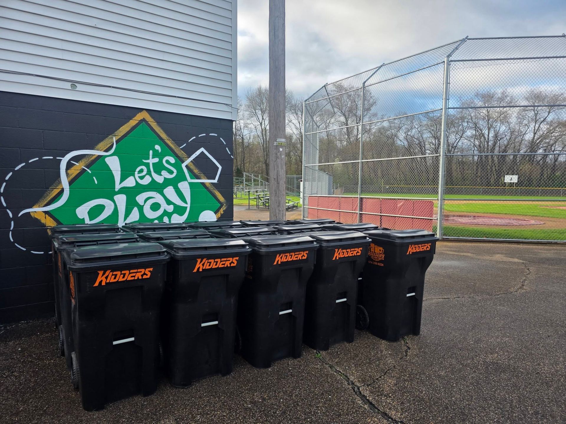Five black recycling bins by a mural reading “Let’s play” beside an outdoor sports court