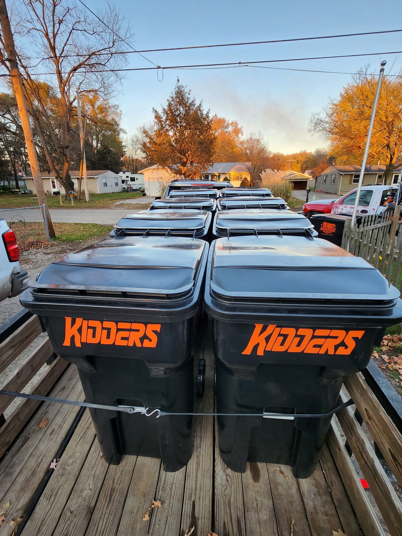 Black KIDDERS trash bins lined up on a truck bed in a residential street at sunset
