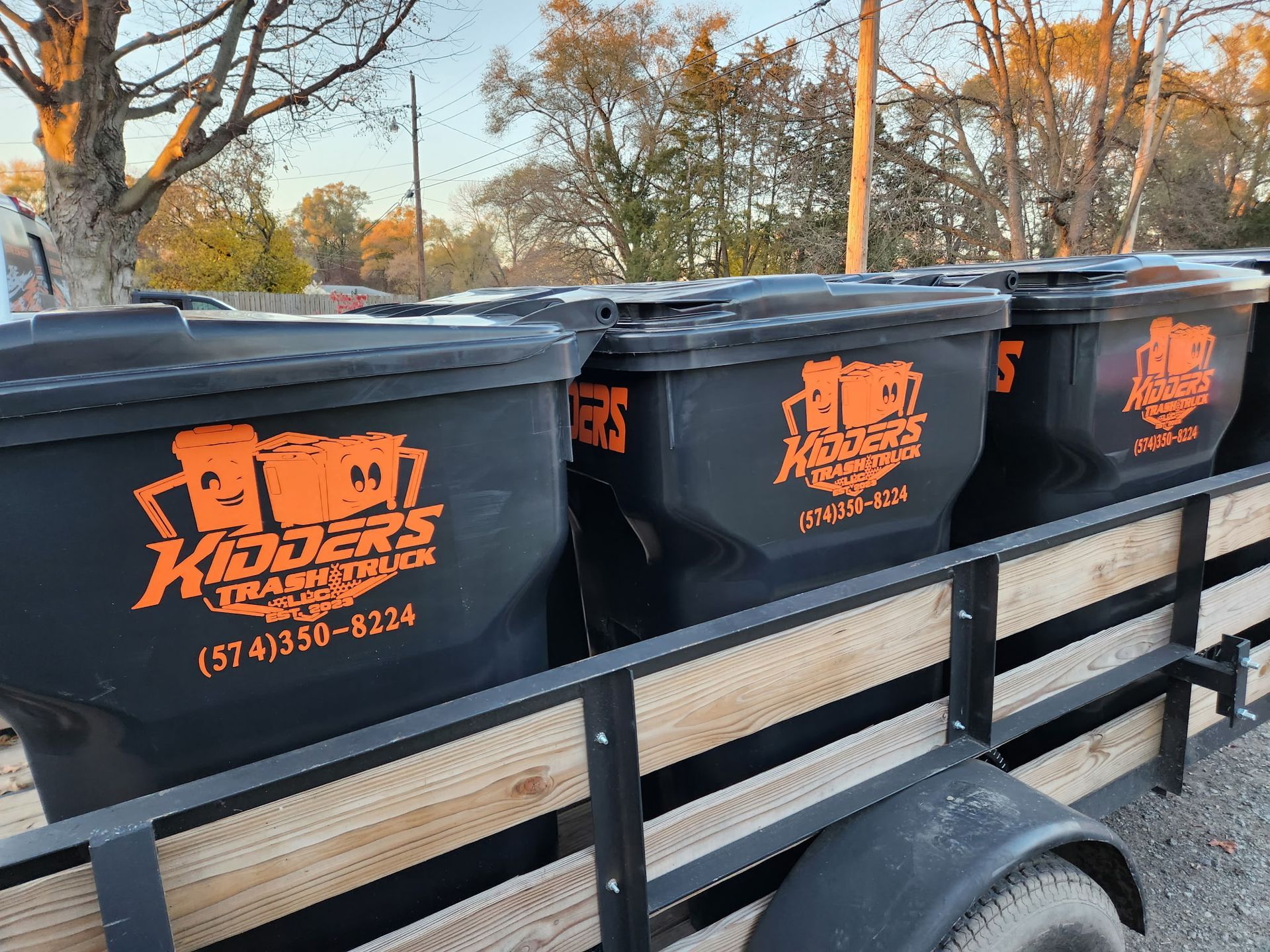 Black trash bins with orange logos lined up on a trailer outdoors in fall.