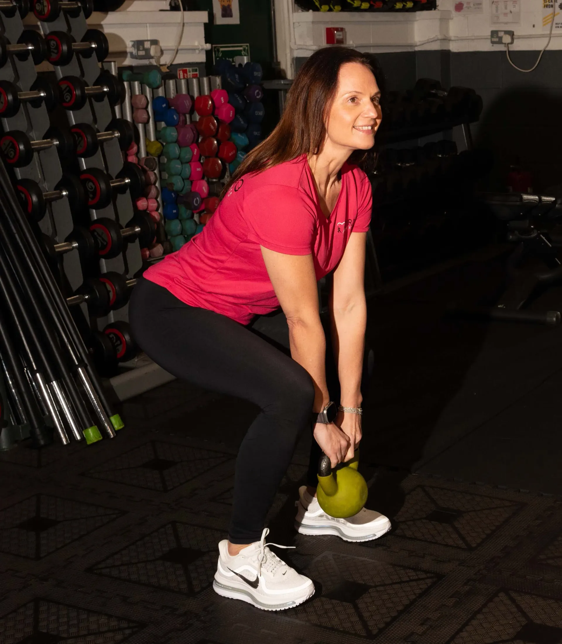 A person in a pink shirt and black leggings performs a kettlebell deadlift in a gym, holding a green kettlebell.